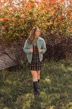 Load image into Gallery viewer, A woman stands outdoors in a grassy area with autumn foliage, wearing a sage green cardigan over a dark plaid dress and black knee-high boots.