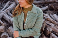 Load image into Gallery viewer, A woman wearing a light sage green chunky knit button-up cardigan over an olive green turtleneck, standing in front of a pile of cut logs.