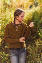 Load image into Gallery viewer, A woman with her hair in a braid stands in a sunlit outdoor field wearing a textured olive-brown knit sweater, gray jeans, and layered necklaces.