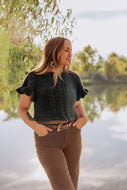 A woman wearing a dark green cable-knit short-sleeve top and brown pants stands outdoors by a calm lake with trees in the background.