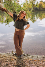 Load image into Gallery viewer, A woman stands outdoors by a calm lake, leaning against a tree branch. She is wearing a dark green textured knit short-sleeve top, brown wide-leg pants, and dark boots.