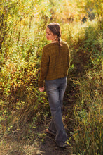 Load image into Gallery viewer, A woman with a long braid walks away on a dirt path surrounded by tall grass and trees, wearing a textured olive-brown cable-knit sweater and blue jeans.