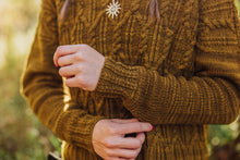 Load image into Gallery viewer, A close-up of a person wearing a chunky cable-knit sweater in a warm mustard brown color, featuring a small white sun-shaped brooch on the chest, with a soft-focus outdoor background.