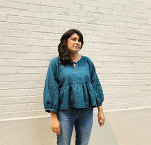 Load image into Gallery viewer, Woman wearing a teal blouse and blue jeans standing against a light gray brick wall.