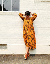 Load image into Gallery viewer, Woman wearing a long orange dress with black patterns against a white brick wall.