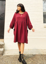 Load image into Gallery viewer, Woman wearing a burgundy dress with long sleeves against a white wall.
