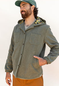 Man wearing a green jacket with a hood and camouflage lining, standing against a plain background.
