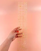Load image into Gallery viewer, Hand holding a clear ruler with gold grid lines against a pink background