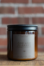 Load image into Gallery viewer, An amber glass jar candle with a black metal lid and a beige paper label reading 'FOG AND FLANNEL' by Seventh House Candles, set on a wooden surface with a blurred brick wall background.