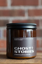 Load image into Gallery viewer, A dark amber glass jar candle with a black lid and a dark brown label reading 'GHOST STORIES' and 'Marshmallow &amp; Campfire' by Seventh House Flame &amp; Wax Co., set on a wooden surface with a blurred brick wall background.
