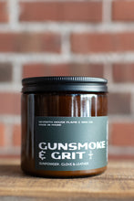 Load image into Gallery viewer, A glass jar candle labeled 'GUNSMOKE &amp; GRIT' by Seventh House Flame &amp; Wax Co., set on a wooden surface with a blurred red brick wall background.