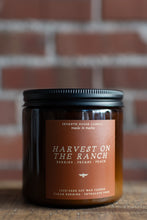 Load image into Gallery viewer, A Seventh House Candle jar candle labeled 'Harvest on the Ranch' in an amber glass container with a black lid, set on a wooden surface against a blurred red brick wall background.