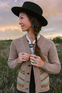 Woman wearing a brown cardigan with a patterned design, standing in a field with a sunset sky.