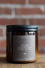 Load image into Gallery viewer, An amber glass jar candle with a black metal lid and a dark taupe label reading 'HIGH COUNTRY CASHMERE - PATCHOULI' by Seventh House Candle, set on a wooden surface with a blurred red brick background.
