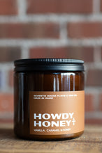 Load image into Gallery viewer, A jar candle labeled 'HOWDY HONEY' with vanilla, caramel, and honey scent, made in Maine by Seventh House Flame &amp; Wax Co., set on a wooden surface with a blurred brick wall background.