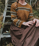 Person wearing a brown sweater with patterned sleeves and a long brown skirt, sitting on a wooden staircase.