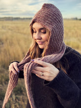 Load image into Gallery viewer, A woman outdoors wearing a chunky ribbed knit bonnet and matching scarf in a dusty mauve-brown color, with a dark knit sweater, against a blurred field background.