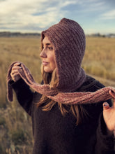 Load image into Gallery viewer, A woman wearing a chunky knit brown hooded beanie and matching scarf, holding the scarf out in an outdoor field.