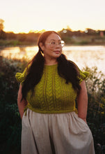 Load image into Gallery viewer, A woman with glasses and long dark hair stands outdoors near a body of water at sunset, wearing a textured olive green knit top with ruffled sleeves and a light beige skirt.