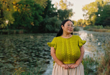 Load image into Gallery viewer, A woman wearing a bright yellow-green cable-knit short-sleeve top and a light beige skirt stands outdoors by a pond with trees in the background.