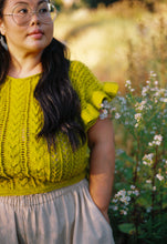 Load image into Gallery viewer, A woman wearing a bright yellow-green cable-knit ruffle-sleeve top and a light beige skirt, standing outdoors in a sunlit field with small white flowers.