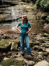Load image into Gallery viewer, A woman stands on mossy rocks in front of a small waterfall and forest stream, wearing a teal short-sleeve knit top and distressed blue jeans.