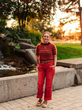 Load image into Gallery viewer, Person wearing a red sweater and pants standing in a park with trees and water features.
