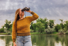 Load image into Gallery viewer, A woman wearing a mustard yellow knit sweater and light blue jeans stands by a lake, looking through black binoculars against a blurred background of trees and a cloudy sky.