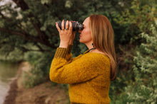 Load image into Gallery viewer, A woman in a textured mustard yellow knit sweater looks through binoculars outdoors against a soft-focus background of green trees and water.