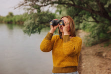 Load image into Gallery viewer, A woman in a textured mustard yellow sweater looks through binoculars outdoors by a river with trees in the background.