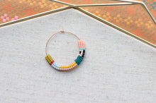 Load image into Gallery viewer, A handmade hoop earring with a gold-tone wire base and a series of colorful seed beads, displayed on a textured light gray fabric surface.