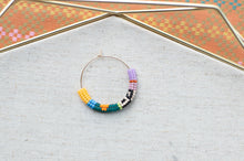 Load image into Gallery viewer, A handmade beaded hoop earring with a gold wire base, decorated with colorful seed beads in yellow, blue, green, black, white, orange, and purple, displayed on a textured light gray fabric surface.