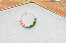Load image into Gallery viewer, A small gold hoop earring adorned with a curved row of colorful seed beads in orange, pink, light blue, teal, and yellow, displayed on a textured beige fabric surface with a geometric gold frame in the background.