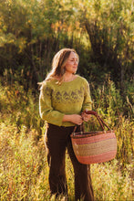 Load image into Gallery viewer, A woman standing in a sunlit grassy field holds a woven basket. She wears a textured olive green sweater and dark brown pants, with a soft-focus background of green and yellow autumn foliage.
