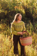 A woman standing in a sunlit grassy field holds a woven basket. She wears a textured olive green sweater and dark brown pants, with a soft-focus background of green and yellow autumn foliage.
