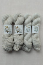 Load image into Gallery viewer, Four skeins of yarn from Pishkun Handwoven Yarn arranged side by side on a textured beige fabric background. The yarn comes in two shades: a warm oatmeal beige on the left and a soft off-white/cream on the right.