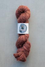 Load image into Gallery viewer, A skein of fluffy, twisted yarn in a warm rust-brown color, featuring a white paper label with a blue and black illustration of a cat and the text 'Pup Paws'. The yarn is photographed on a neutral gray background.