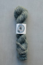 Load image into Gallery viewer, A skein of fuzzy yarn in a muted blue-gray color, wrapped with a white paper label that reads 'Moonbeam'. The yarn is photographed vertically against a neutral gray textured background.