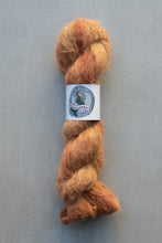 Load image into Gallery viewer, A skein of fluffy, twisted wool or yarn in a warm, mottled orange-brown shade, secured with a white paper label featuring a blue circular design. The product is photographed vertically against a textured, cool gray-blue background.
