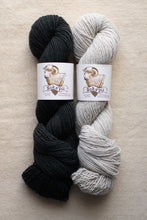 Load image into Gallery viewer, Two skeins of black and gray yarn with labels on a beige background