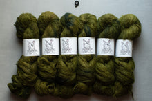 Load image into Gallery viewer, Six skeins of thick, fuzzy yarn in a mottled olive green and dark forest green colorway, arranged in two rows on a light gray background. Each skein is wrapped with a white paper label featuring a black line-art illustration of a dog.