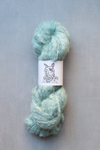 Load image into Gallery viewer, A skein of fluffy, soft yarn in a light mint green or pale aqua color, wrapped with a white paper label featuring a black line drawing of a llama and the text 'Old Darn Dog'. The yarn is photographed on a neutral, textured gray background.