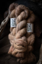 Load image into Gallery viewer, A close-up of a thick braided bundle of soft, fuzzy yarn or fiber in warm taupe and light brown tones, with two white paper labels wrapped around it. The background is very dark and moody, with rustic wood elements visible at the edges.