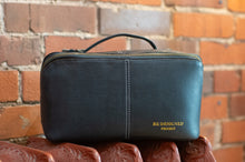 Load image into Gallery viewer, A dark charcoal gray leather cosmetic pouch with gold lettering that reads 'BE-DESIGNED PROJECT' on the front, resting on a carved wooden surface against a brick wall background.