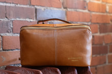 Load image into Gallery viewer, Brown leather toiletry bag with 'Redesigned' branding against a brick wall.