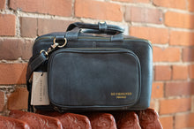 Load image into Gallery viewer, A dark gray leather briefcase-style bag with gold 'REDESIGNED PROJECT' branding, resting on a carved wooden surface against a red brick wall background.