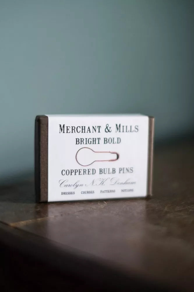 Box of Merchant & Mills copper bulb pins on a wooden surface with a dark background