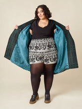 Load image into Gallery viewer, Woman wearing a black top, patterned shorts, and holding a green jacket against a beige background