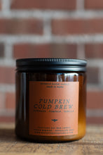 Load image into Gallery viewer, An amber glass jar candle with a black metal lid and a terracotta-colored label reading 'Pumpkin Cold Brew' by Seventh House Candles, set on a wooden surface with a blurred brick wall background.