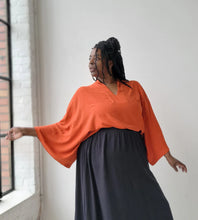 Load image into Gallery viewer, Woman wearing an orange top and black skirt standing by a window with white walls.
